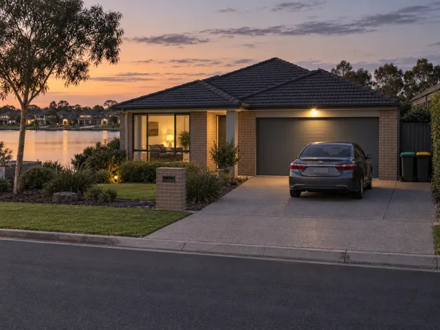 A brick lakeside house at dusk with a lamp lit in the front room, a light on above the garage door, a car in the driveway and two wheelie bins beside the side fence